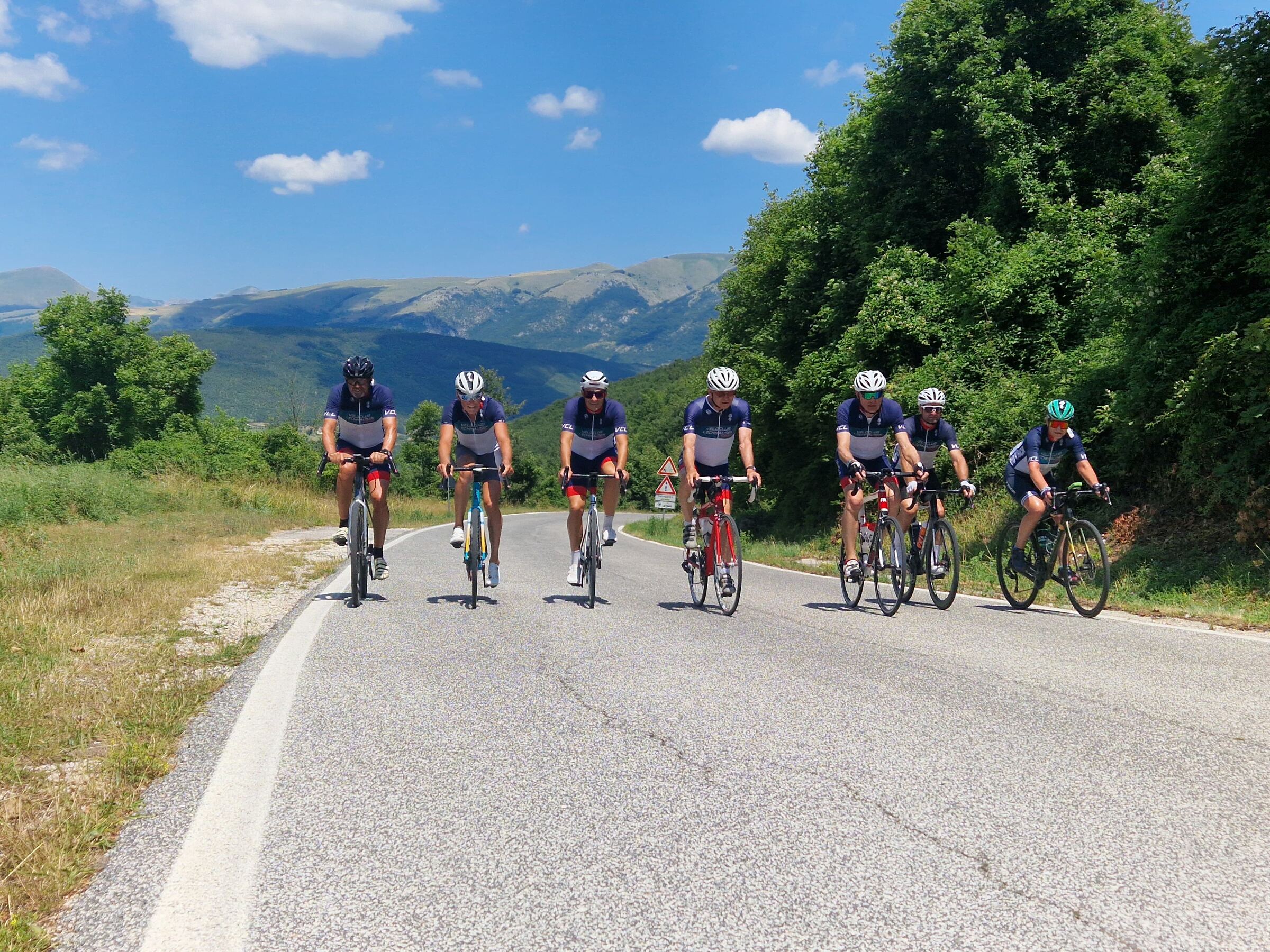 Sieben Mitglieder des Veloclub Lechhausen fahren im Vereinstrikot nebeneinander auf einer Bergstraße, im Hintergrund Berge und blauer Himmel